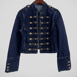 A dark blue denim jacket with decorative gold-colored military-style buttons, shown hanging on a wooden hanger against a white wall.
