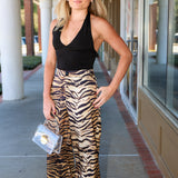 A woman posing outdoors on a covered walkway, wearing a black halter top and high-waisted, wide-leg trousers with a brown and black tiger stripe pattern. She is holding a small clear handbag.