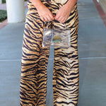 A person wearing high-waisted, wide-leg trousers with a bold black and tan tiger stripe pattern, holding a clear handbag on a concrete walkway.