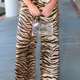 A person wearing high-waisted, wide-leg trousers with a bold black and tan tiger stripe pattern, holding a clear handbag on a concrete walkway.