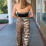 A woman viewed from behind wearing a strapless black bandeau top and high-waisted wide-leg trousers with a beige, brown, and black tiger stripe pattern, standing on a covered outdoor walkway.