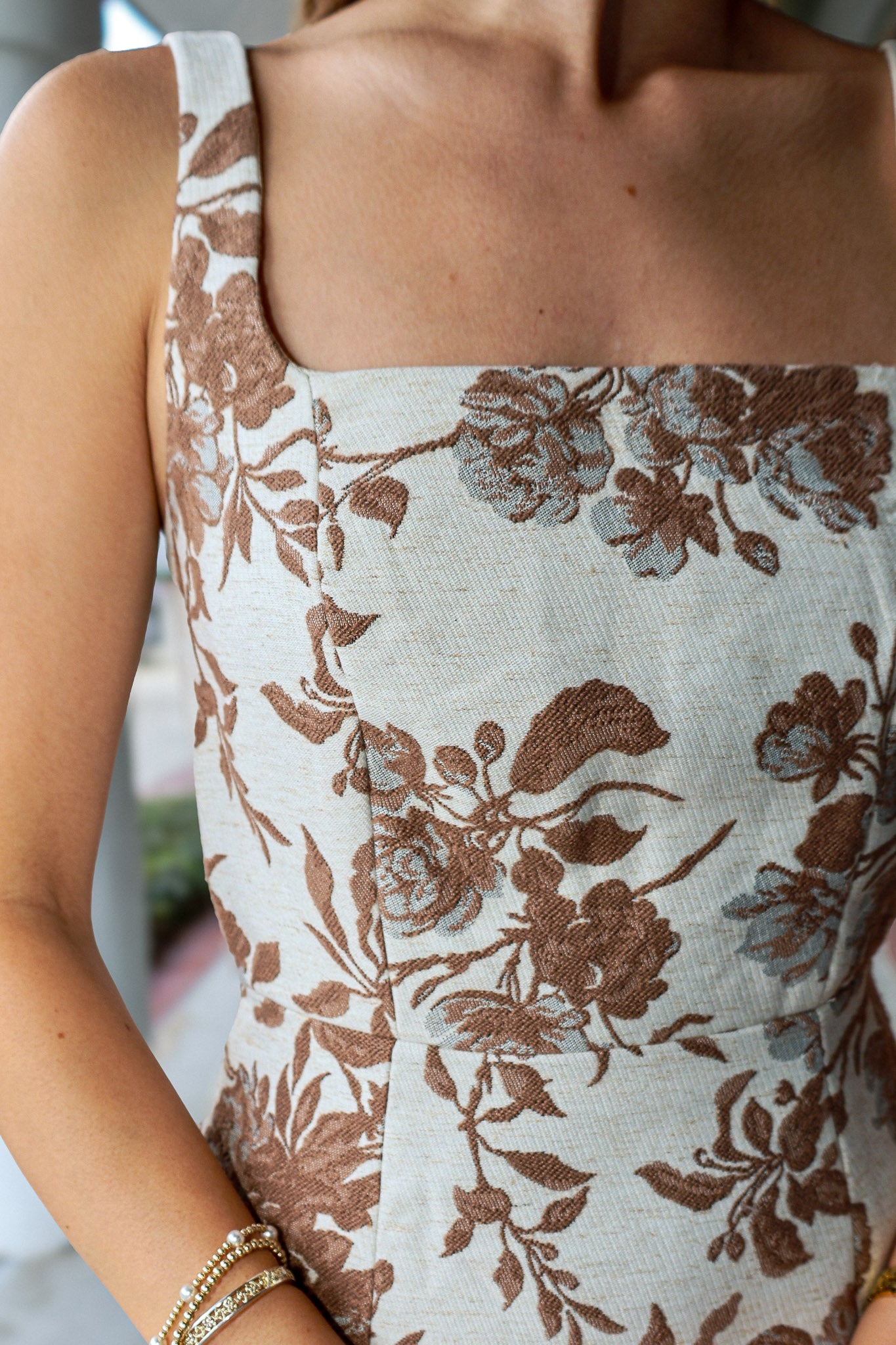 Close-up of a woman wearing a sleeveless top with a square neckline and a brown floral jacquard pattern on a light beige fabric.