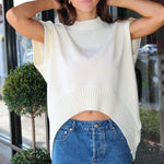 A woman wearing a cream-colored, sleeveless ribbed knit top with a curved hem and a blue denim mini skirt, posing in front of a glass storefront.