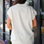 A woman viewed from behind wearing an oversized cream-colored knit sweater vest with short sleeves and ribbed trim, standing in front of a glass storefront.