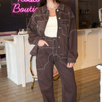 A woman wearing a dark brown western-style denim jacket and matching dark brown jeans with light contrast stitching, posing in front of a pink 'Rodéo Boutique' neon sign.