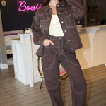 A woman wearing in a brown denim jacket and matching brown denim pants with white contrast stitching, styled with a cream tank top and white sneakers at Rodeo Boutique.