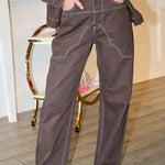 A woman wearing a dark brown western-style denim jacket and matching dark brown jeans with light contrast stitching, posing in front of a pink 'Rodéo Boutique' neon sign.