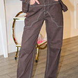A woman wearing a dark brown western-style denim jacket and matching dark brown jeans with light contrast stitching, posing in front of a pink 'Rodéo Boutique' neon sign.