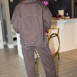 A woman wearing a dark brown western-style denim jacket and matching dark brown jeans with light contrast stitching, posing in front of a pink 'Rodéo Boutique' neon sign.