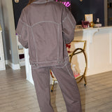 A woman wearing a dark brown western-style denim jacket and matching dark brown jeans with light contrast stitching, posing in front of a pink 'Rodéo Boutique' neon sign.
