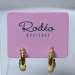 A pair of gold hoop earrings with small clear crystal accents, displayed on a pink card labeled 'Rodeo Boutique'.
