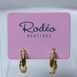 A pair of gold hoop earrings with small clear crystal accents, displayed on a pink card labeled 'Rodeo Boutique'.