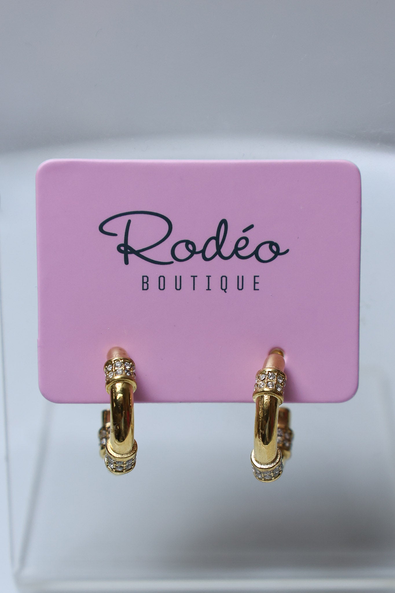 A pair of gold hoop earrings with small clear crystal accents, displayed on a pink card labeled 'Rodeo Boutique'.