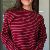 A woman wearing a long-sleeve henley top with horizontal burgundy, white, and dark gray stripes, paired with white pants, captured in a lifestyle setting at Rodeo Boutique.
