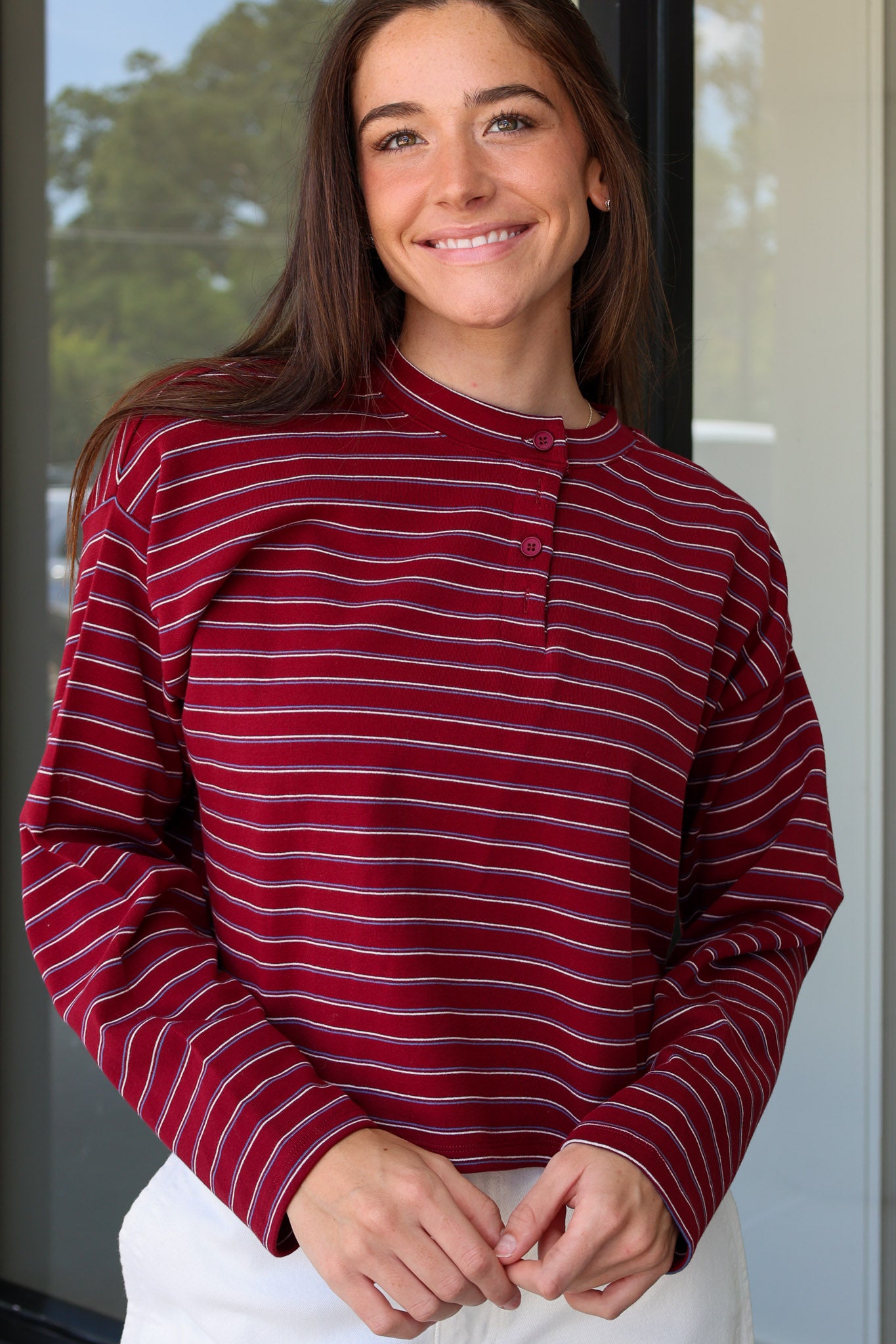 A woman wearing a long-sleeve henley top with horizontal burgundy, white, and dark gray stripes, paired with white pants, captured in a lifestyle setting at Rodeo Boutique.