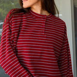 A woman wearing a long-sleeve henley top with horizontal burgundy, white, and dark gray stripes, paired with white pants, captured in a lifestyle setting at Rodeo Boutique.