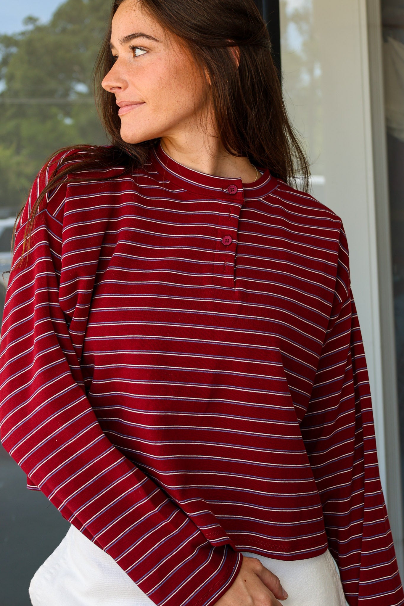 A woman wearing a long-sleeve henley top with horizontal burgundy, white, and dark gray stripes, paired with white pants, captured in a lifestyle setting at Rodeo Boutique.
