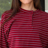 A woman wearing a long-sleeve henley top with horizontal burgundy, white, and dark gray stripes, paired with white pants, captured in a lifestyle setting at Rodeo Boutique.