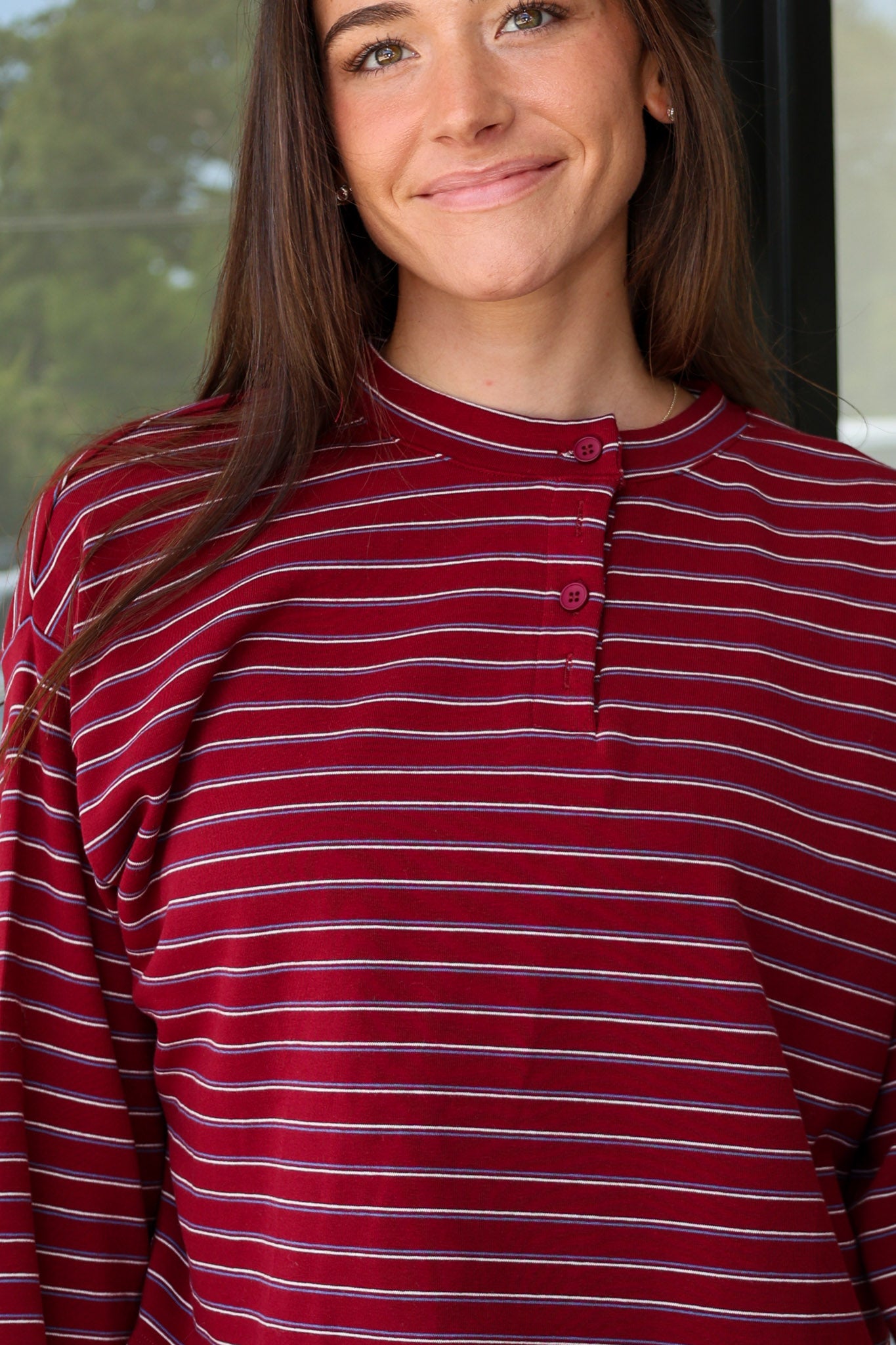 A woman wearing a long-sleeve henley top with horizontal burgundy, white, and dark gray stripes, paired with white pants, captured in a lifestyle setting at Rodeo Boutique.