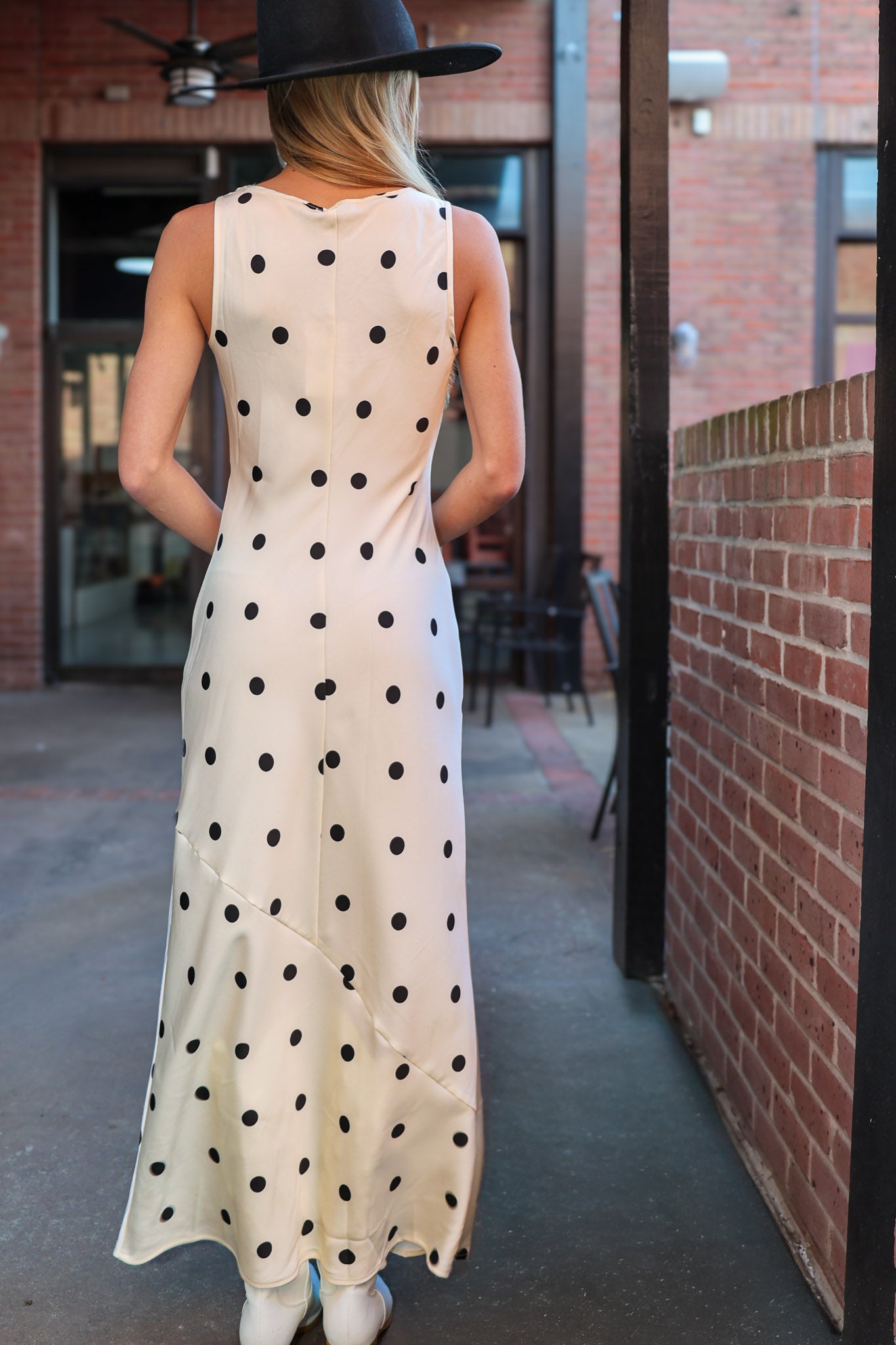 Woman wearing a polka dot dress and black hat walking away from the camera.
