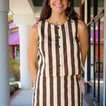 A woman smiling while wearing a sleeveless brown and cream vertical striped button-front top and matching mini skirt set, standing in an outdoor covered walkway.