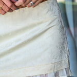 Close-up of a person wearing a short cream or off-white skirt with a subtle tonal floral jacquard pattern and a ruffled hem. The person is also wearing a dark brown top and several rings.