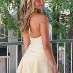 A woman wearing a pale yellow sleeveless mini dress with thin straps and a ruffled hem, photographed outdoors on a balcony with trees and a house in the background.