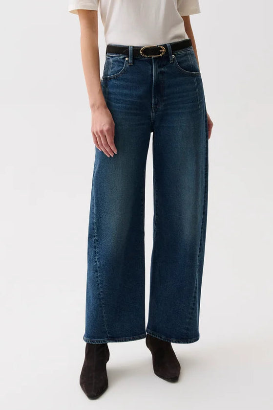 A woman modeling a pair of dark blue wide-leg jeans with a black belt, paired with a cream short-sleeve t-shirt and dark brown ankle boots, against a plain white studio background.