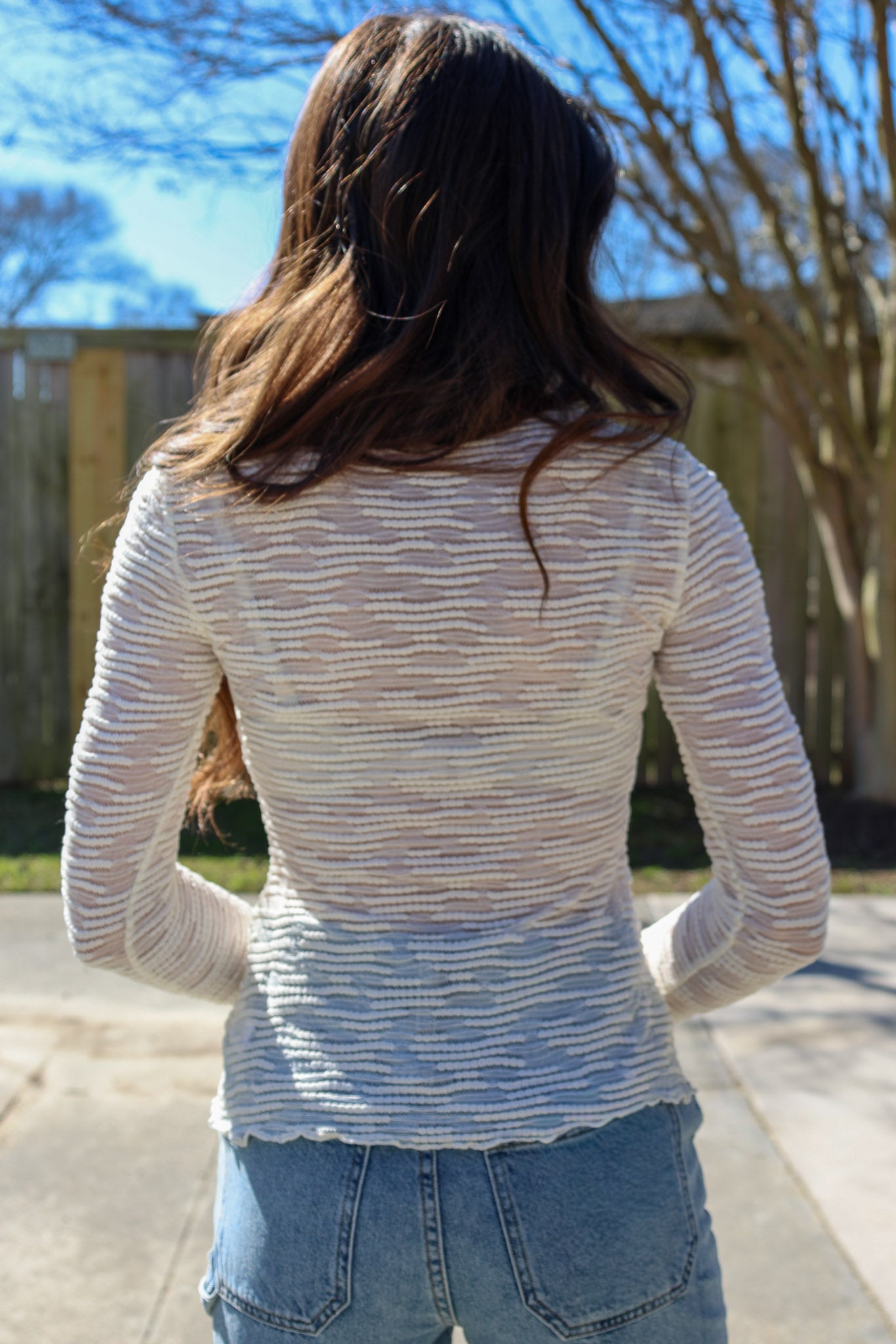 Person wearing a light-colored sheer sweater and jeans standing outdoors with trees and blue sky in the background
