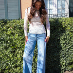 Woman wearing a light-colored sheer top and blue jeans standing in front of a green hedge.