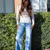 Woman wearing a light-colored sheer top and blue jeans standing in front of a green hedge.