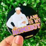 A hand holds a round black button badge featuring a stylized illustration of a person in a white hoodie and visor, with the text 'LANE IS MY HOMEBOY' in yellow and purple. The background is a blurred green hedge.