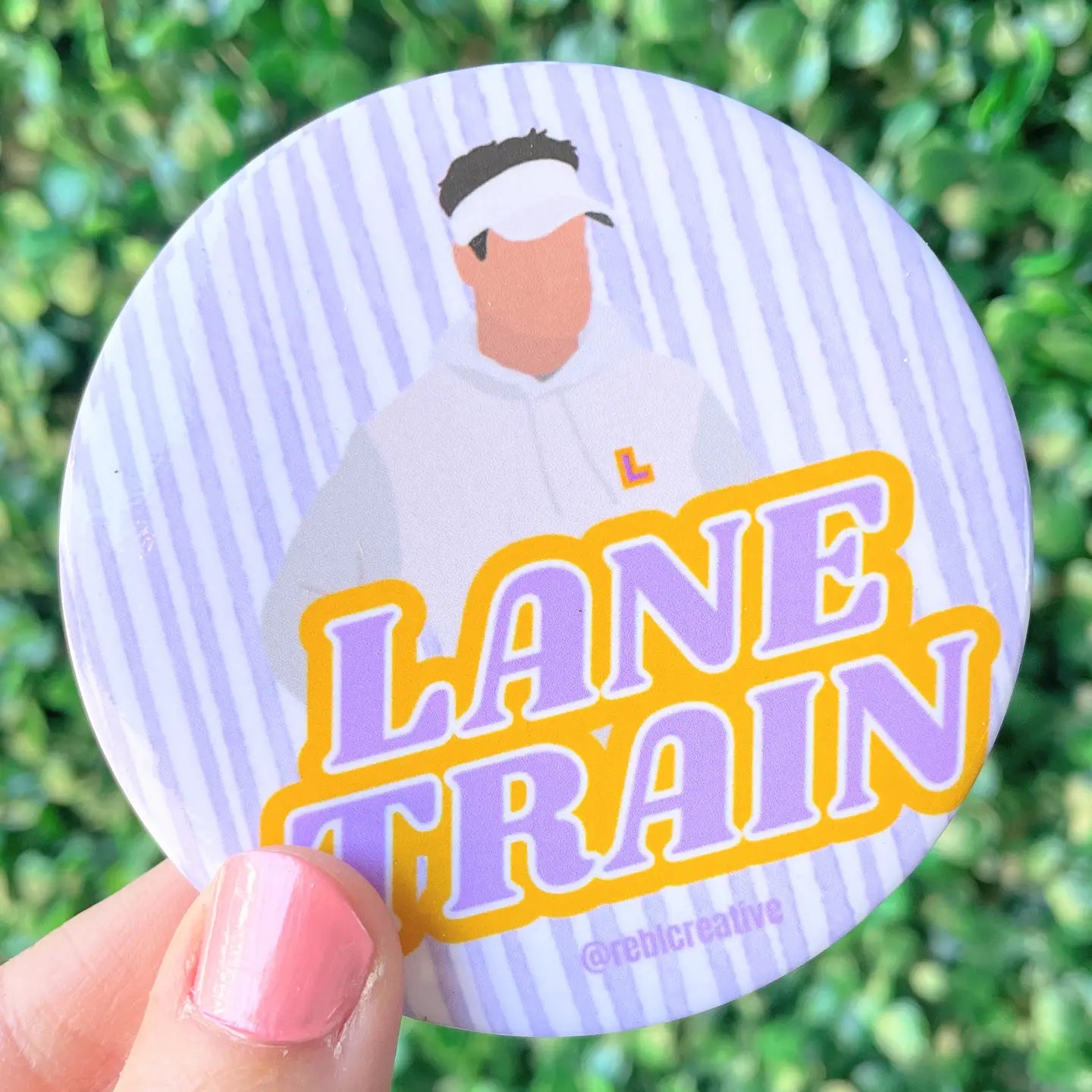 A round pinback button badge held by a hand, featuring a stylized illustration of a man in a white hoodie and visor on a light purple and white striped background, with the text 'LANE TRAIN' in purple and yellow. The background is a blurred green hedge.