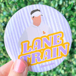 A round pinback button badge held by a hand, featuring a stylized illustration of a man in a white hoodie and visor on a light purple and white striped background, with the text 'LANE TRAIN' in purple and yellow. The background is a blurred green hedge.