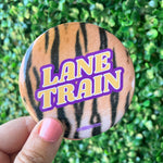 A round pinback button badge with a tiger-stripe pattern and the words 'LANE TRAIN' in bold yellow letters with a purple outline, held by a hand against a green hedge background.