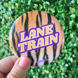 A round pinback button badge with a tiger-stripe pattern and the words 'LANE TRAIN' in bold yellow letters with a purple outline, held by a hand against a green hedge background.