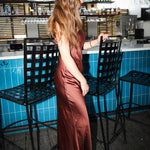 A woman with long light brown hair stands in a bar setting wearing a floor-length satin slip dress in a warm rust or terracotta brown color. She is positioned in front of a blue-tiled counter and black metal bar stools.