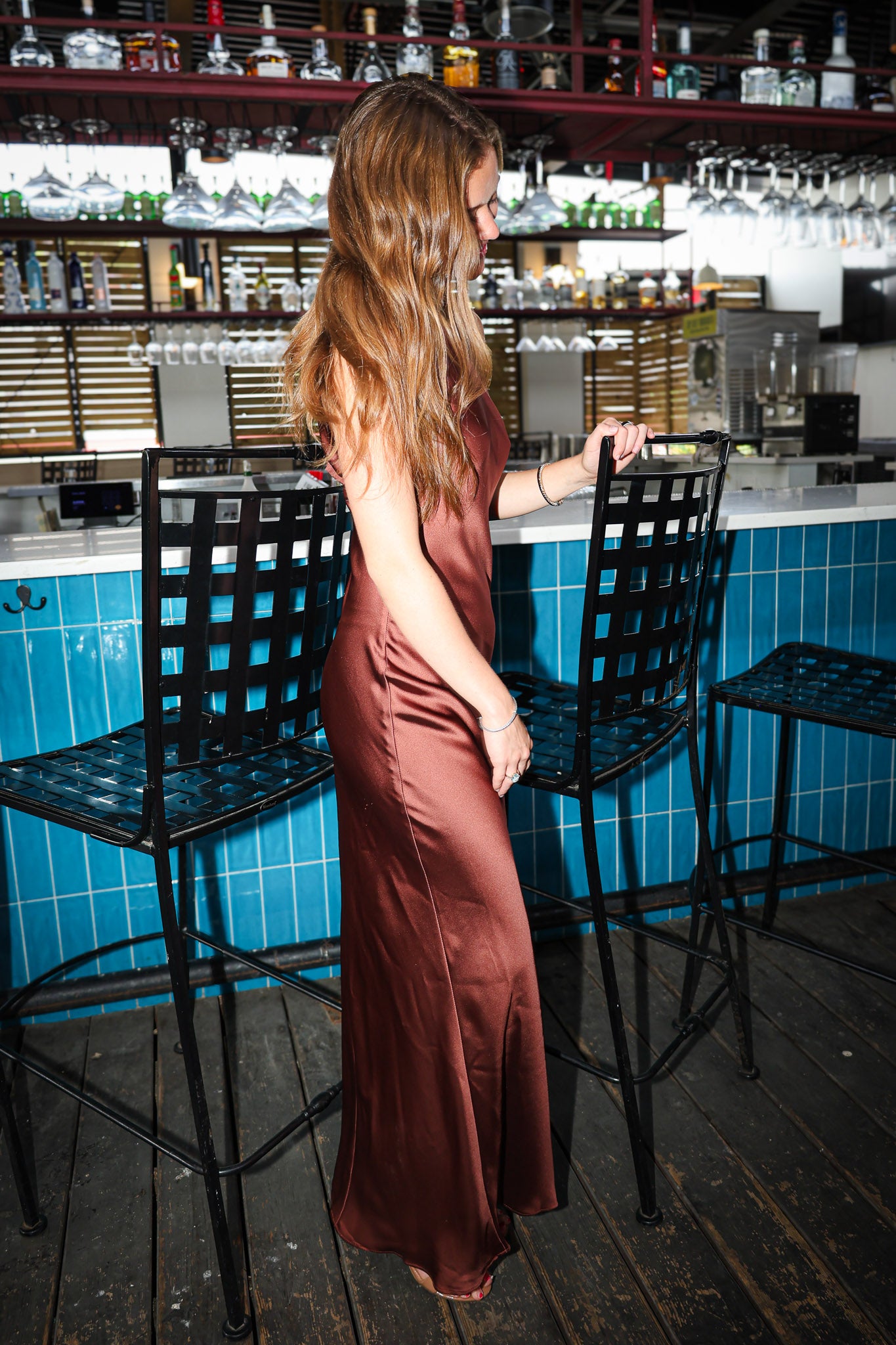 A woman with long light brown hair stands in a bar setting wearing a floor-length satin slip dress in a warm rust or terracotta brown color. She is positioned in front of a blue-tiled counter and black metal bar stools.
