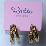 A pair of gold hoop earrings with a twisted, rope-like textured design, displayed on a pink card labeled 'Rodéo BOUTIQUE' against a light gray background.