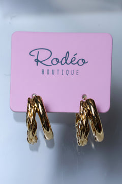 A pair of gold hoop earrings with a twisted, rope-like textured design, displayed on a pink card labeled 'Rodéo BOUTIQUE' against a light gray background.