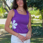 A woman standing outdoors at LSU wearing a purple sleeveless top with white flower graphics and a white mini skirt.
