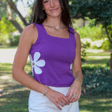 A woman standing outdoors at LSU wearing a purple sleeveless top with white flower graphics and a white mini skirt.