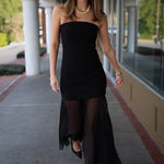 Woman wearing a strapless black dress with sheer overlay on a sidewalk.