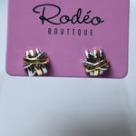 A pair of small wrap-style earrings displayed on a pink card labeled 'Rodéo Boutique.' The earrings appear metallic with a mix of gold, silver, and possibly rose-gold tones, set against a soft light gray background.