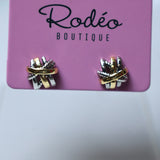 A pair of small wrap-style earrings displayed on a pink card labeled 'Rodéo Boutique.' The earrings appear metallic with a mix of gold, silver, and possibly rose-gold tones, set against a soft light gray background.