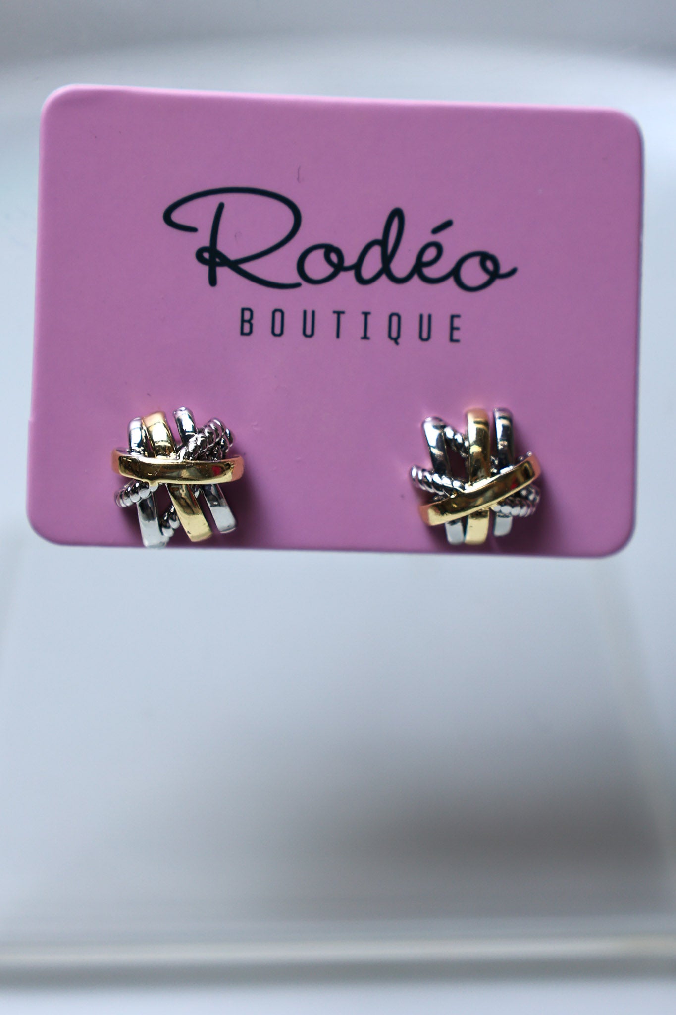 A pair of small wrap-style earrings displayed on a pink card labeled 'Rodéo Boutique.' The earrings appear metallic with a mix of gold, silver, and possibly rose-gold tones, set against a soft light gray background.
