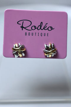 A pair of small wrap-style earrings displayed on a pink card labeled 'Rodéo Boutique.' The earrings appear metallic with a mix of gold, silver, and possibly rose-gold tones, set against a soft light gray background.