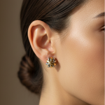 A close-up side profile of a woman wearing a small gold and silver floral stud earring, with her hair pulled back and a white collar visible against a warm taupe background.