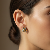 A close-up side profile of a woman wearing a small gold and silver floral stud earring, with her hair pulled back and a white collar visible against a warm taupe background.