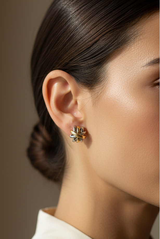 A close-up side profile of a woman wearing a small gold and silver floral stud earring, with her hair pulled back and a white collar visible against a warm taupe background.